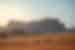 Spire mountain range in distance with two trucks driving past in the distance in an orange sand desert