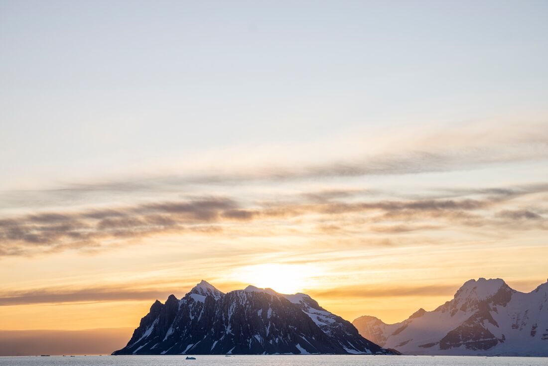 The sunrise emerges above a wide mountain on the Antarctic coast as a zodiac cruises by on the water